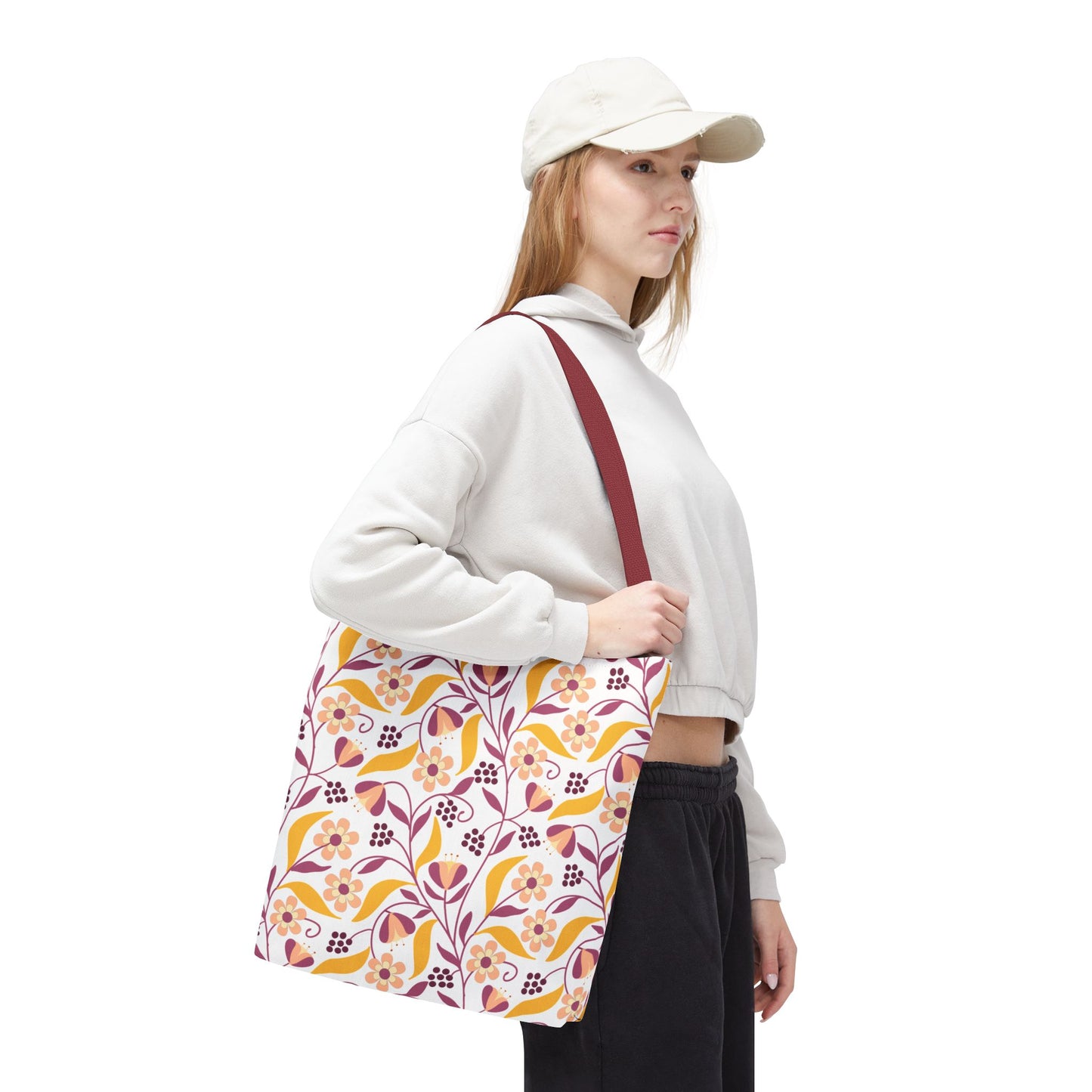 Model wearing a light-beige baseball cap and off-white cropped hoodie holds a Floral Tote Bag with a burgundy strap and cheerful floral pattern, embodying FireBrook Designs' vibrant, joyful style.