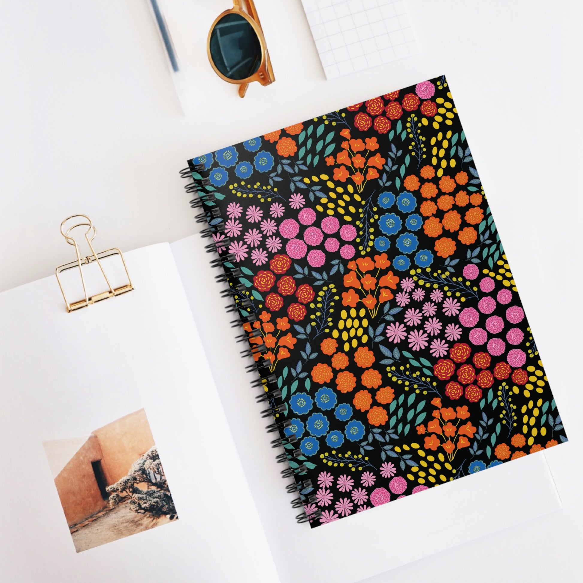 Floral Spiral Notebook with vibrant blooms on a black cover, central in a stylish desk setup, embodying creativity and joy, alongside a magazine and tortoiseshell sunglasses.