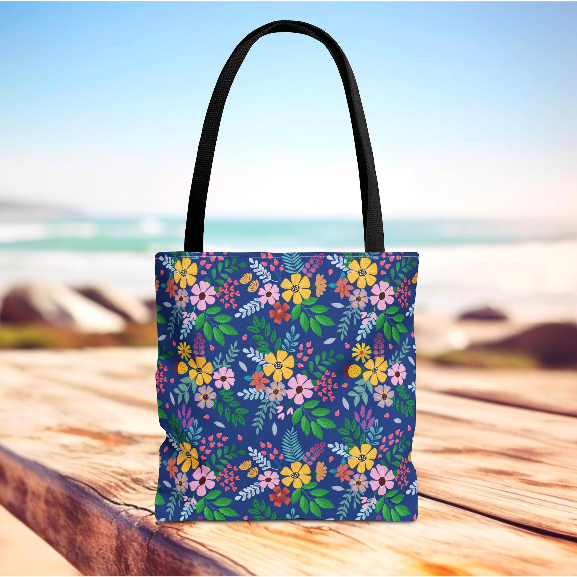 Deep Sea Floral Patch Tote Bag with black straps, featuring vibrant floral patterns on a dark blue background, sits on a wooden surface against a blurred beach backdrop, highlighting its cheerful, summery design.