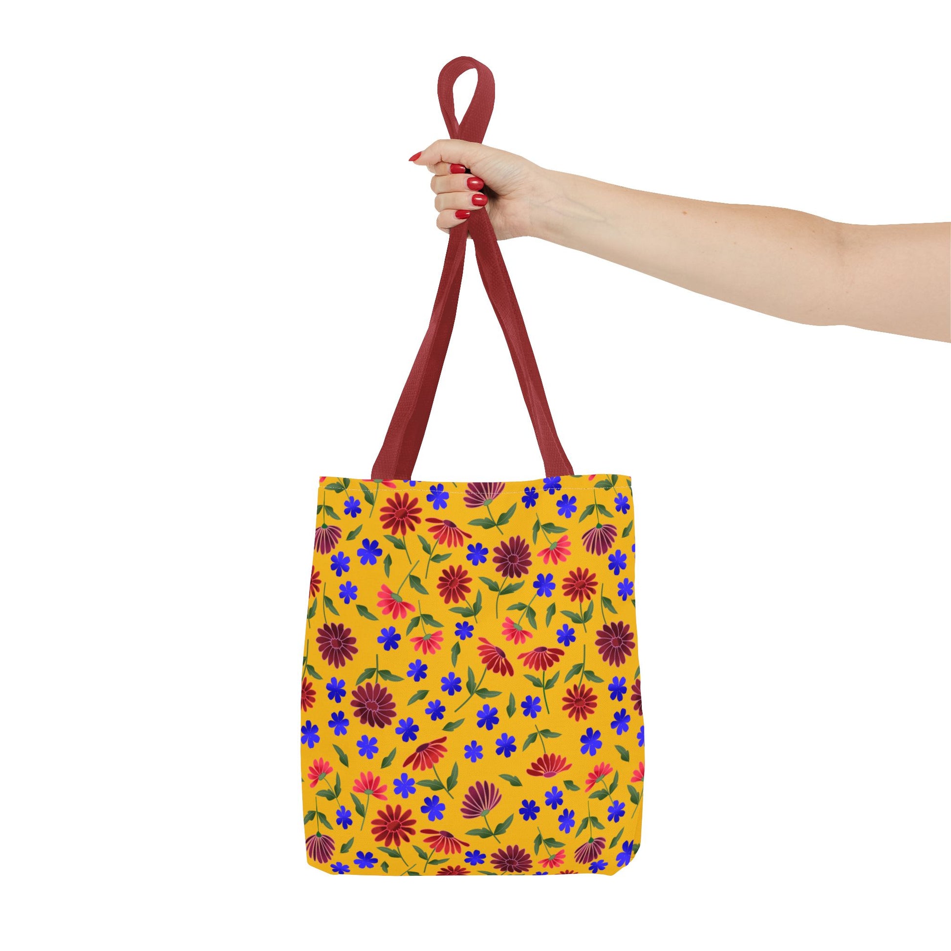 Golden Wildflower Tote Bag: A hand holds a bright yellow tote bag adorned with red daisies, blue flowers, and green leaves, featuring burgundy-red handles, embodying FireBrook Designs’ cheerful, practical style.