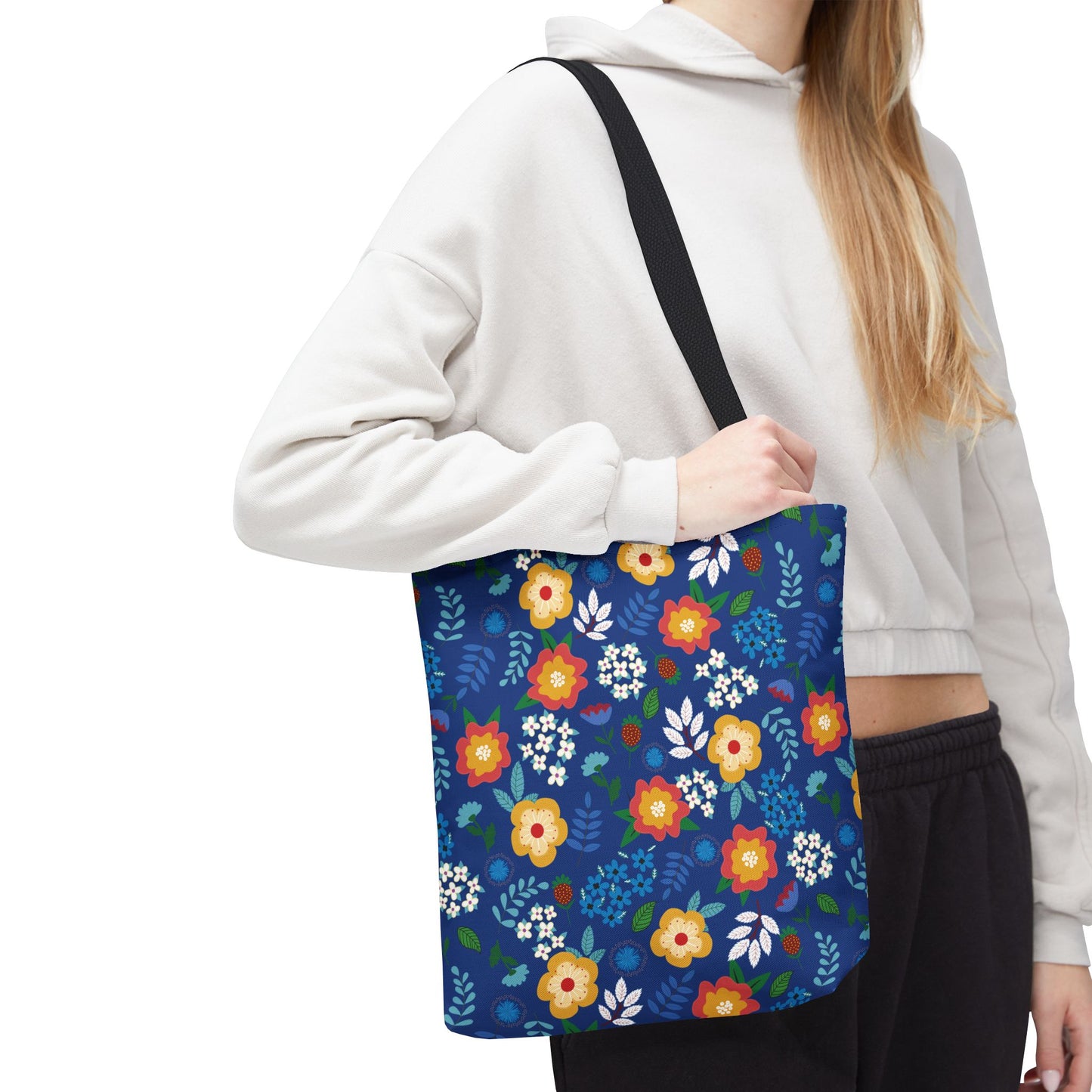 Person wearing a light-colored hoodie carries a Floral Tote Bag, featuring bright blue with yellow, red, white flowers. Ideal for summer outings, this cheerful bag reflects FireBrook Designs' uplifting approach.