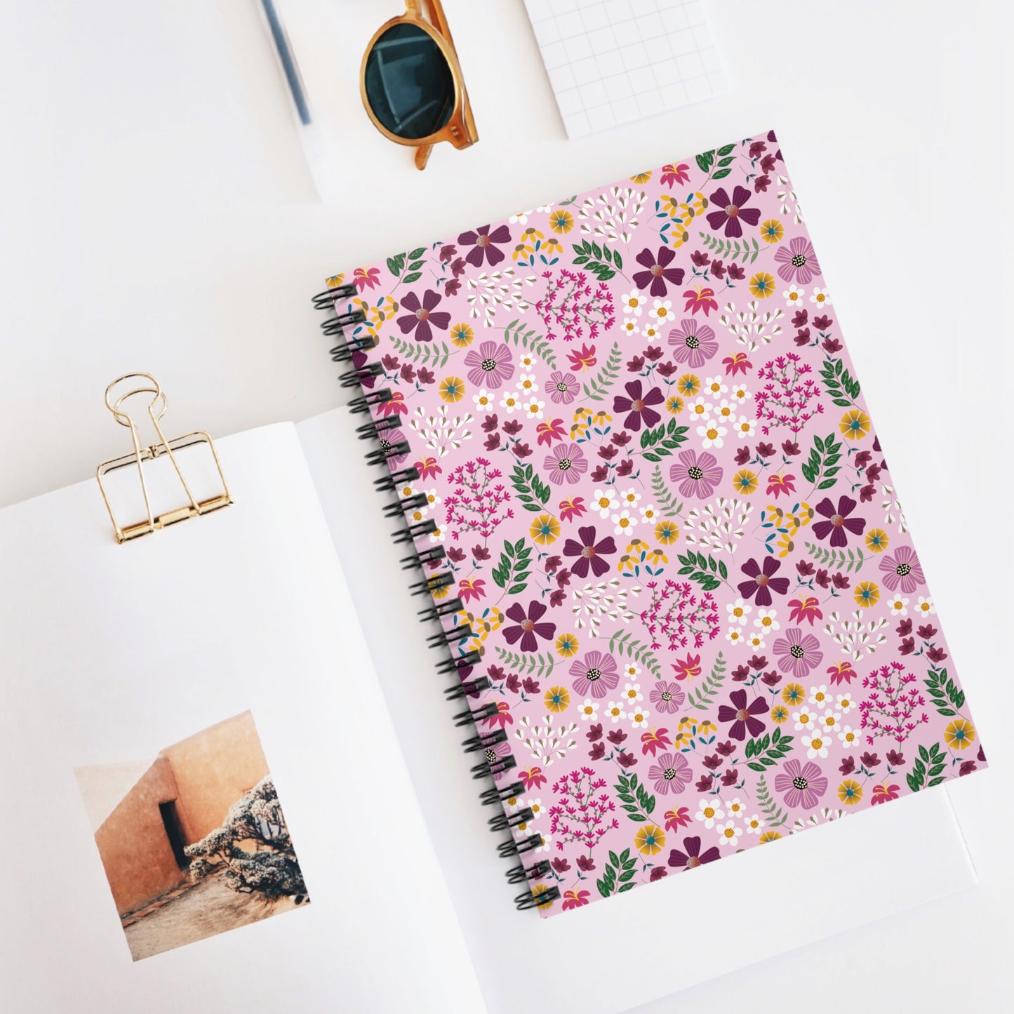 Floral spiral Notebook with vibrant flower patterns lies atop an open book, accompanied by amber sunglasses and grid paper; a joyful, stylish addition to any workspace.