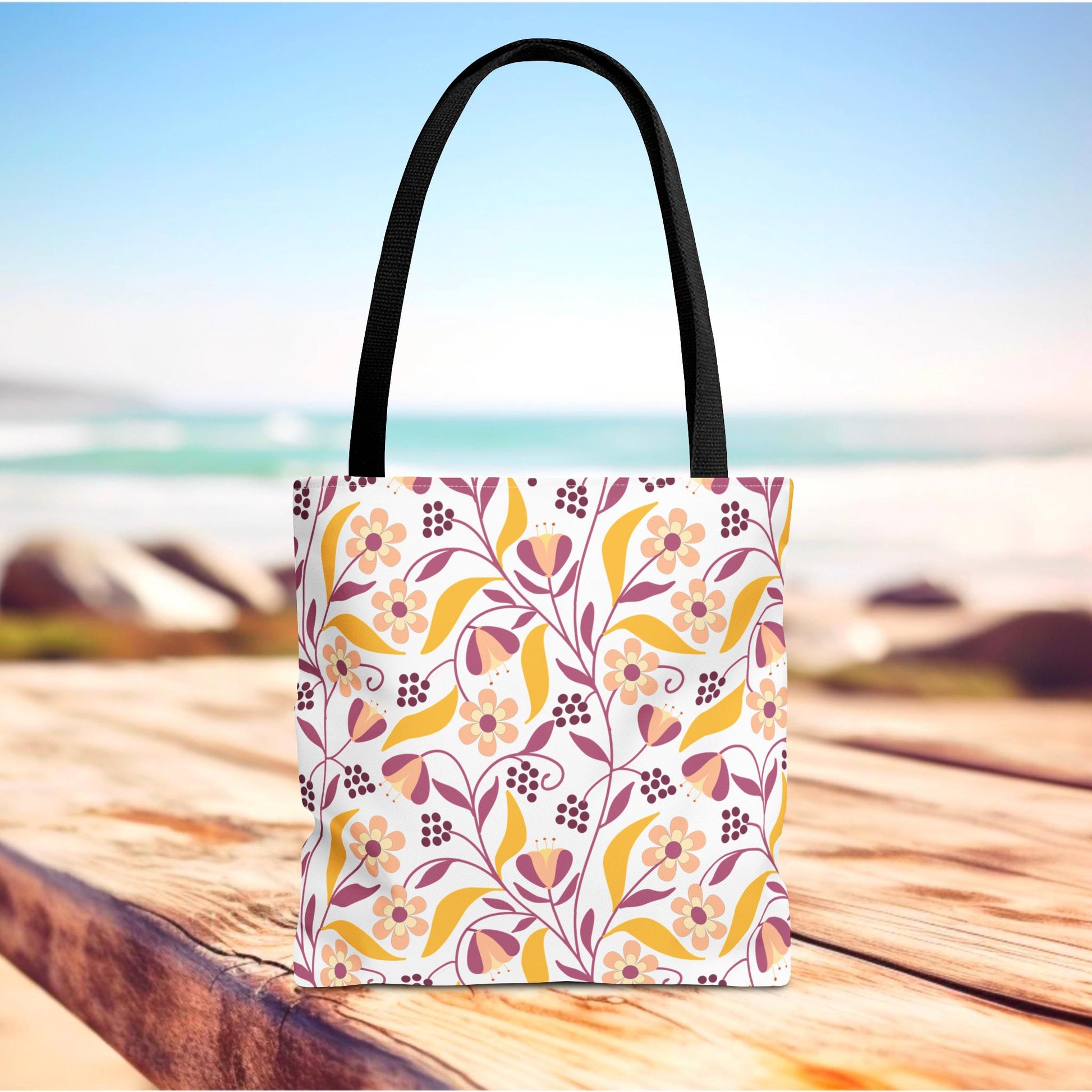 Golden Paisley Floral Tote Bag featuring a soft pink, peach, mustard-yellow, and burgundy floral motif on a white background, sitting on wood near a beach, ideal for joyful summer outings.