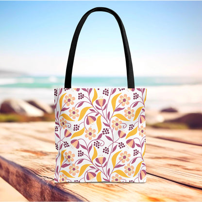 Golden Paisley Floral Tote Bag featuring a soft pink, peach, mustard-yellow, and burgundy floral motif on a white background, sitting on wood near a beach, ideal for joyful summer outings.