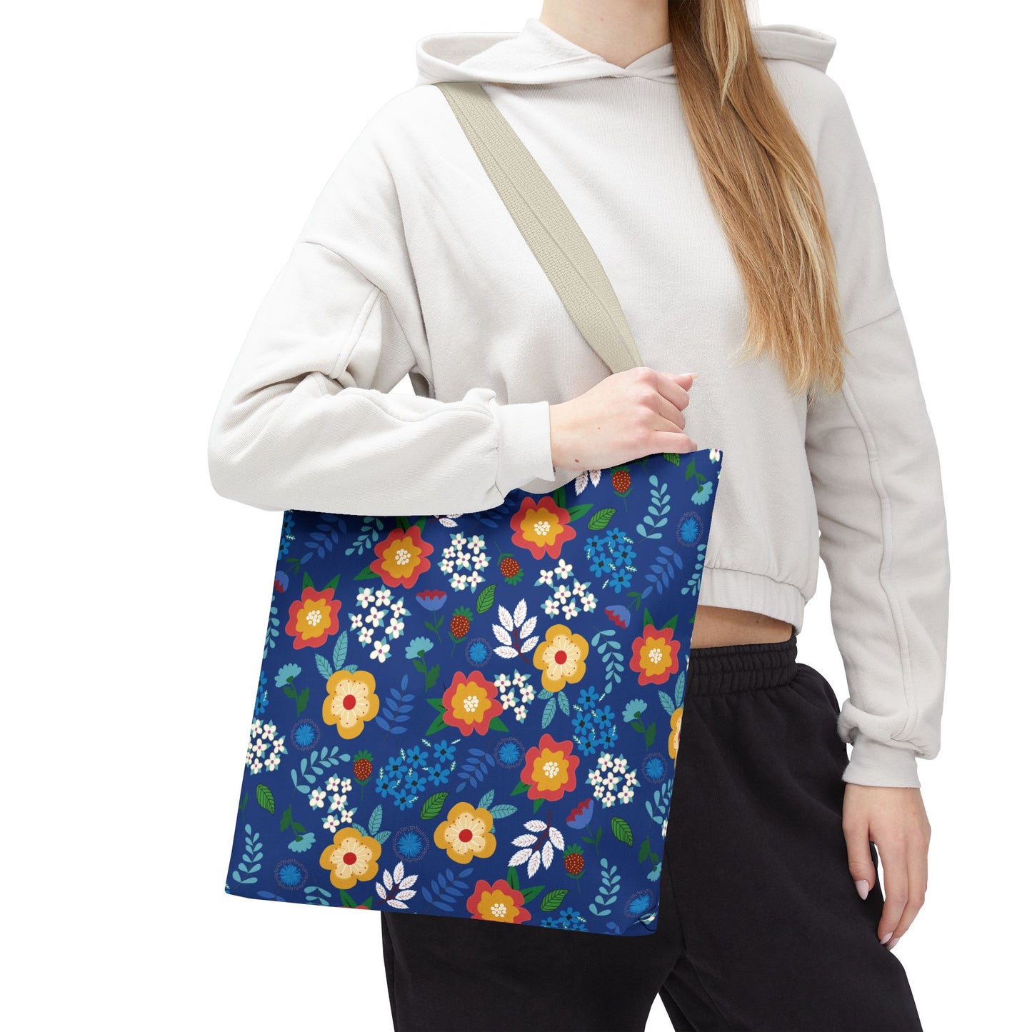 Floral Tote Bag slung over shoulder, showcasing vibrant red, yellow, and blue flowers on a navy background, ideal for summer outings. The person wears a light cropped hoodie and dark pants.