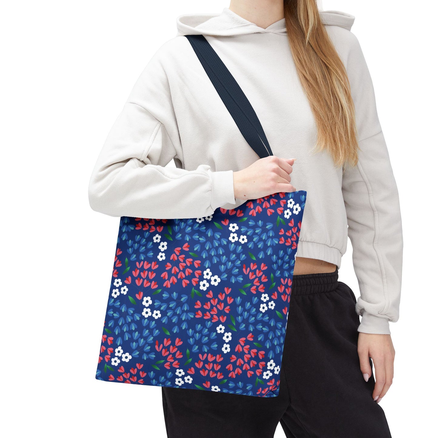 Person wearing a light-colored hoodie and dark pants carries the Floral Tote Bag, featuring a navy-blue canvas with white blossoms and red clusters, embodying FireBrook Designs' cheerful and uplifting aesthetic.