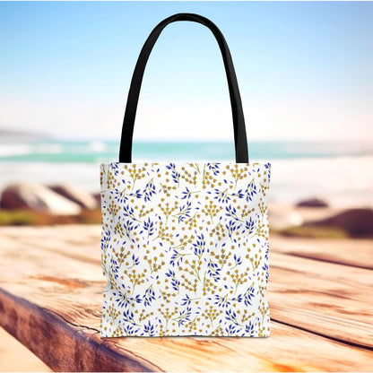 Wild Herb Garden Tote Bag with black straps, featuring a vibrant botanical pattern in blue and mustard on white, stands on a wooden dock against a beach backdrop, ideal for joyful summer outings.