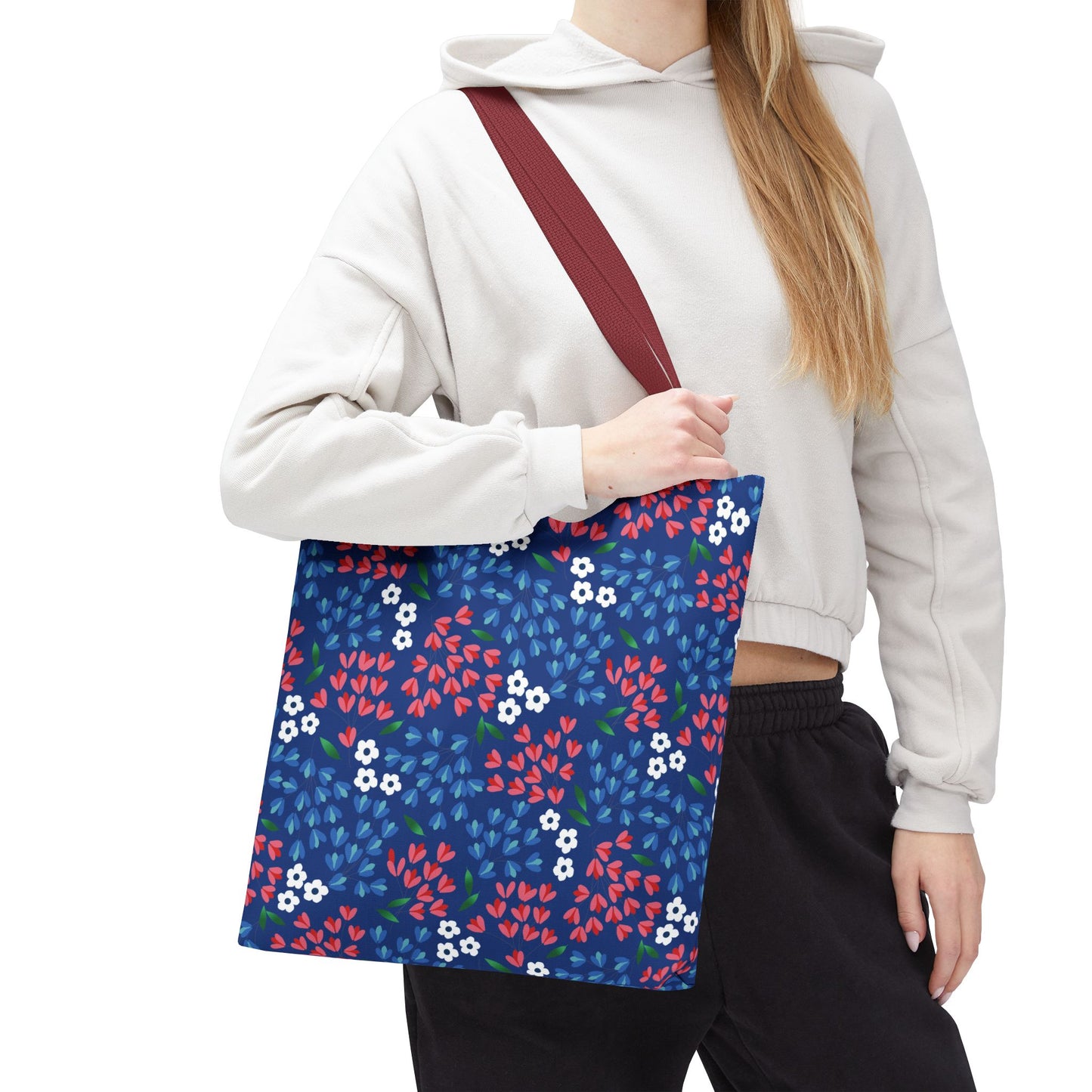 Floral Tote Bag with a navy-blue background featuring red and blue leaf clusters and white blossoms, held by a person wearing a light hoodie. Ideal for summer outings, embraces joyful, stylish design.
