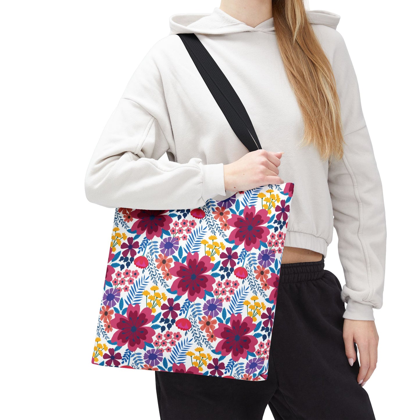 Person wearing a light cropped hoodie and dark pants holds the Violet Daisy Carnival Tote Bag, featuring a vibrant floral pattern with magenta, red, purple, and yellow flowers, showcasing FireBrook Designs' joyful style.