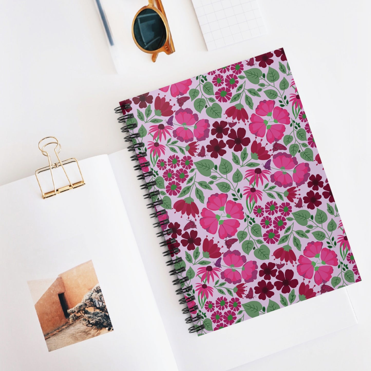 Floral Spiral Notebook with vibrant pink and burgundy flower pattern on a light purple background, surrounded by sunglasses, a small grid notepad, and an open book, reflecting FireBrook Designs' uplifting style.