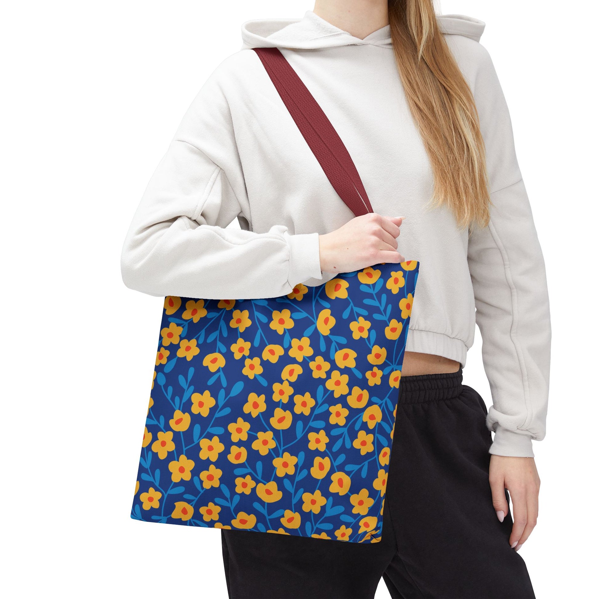 Cobalt Poppy Field Tote Bag with vibrant yellow-orange flowers and blue leaves on a dark blue background, held by a person in a light-gray hoodie, featuring a burgundy strap for stylish, cheerful outings.