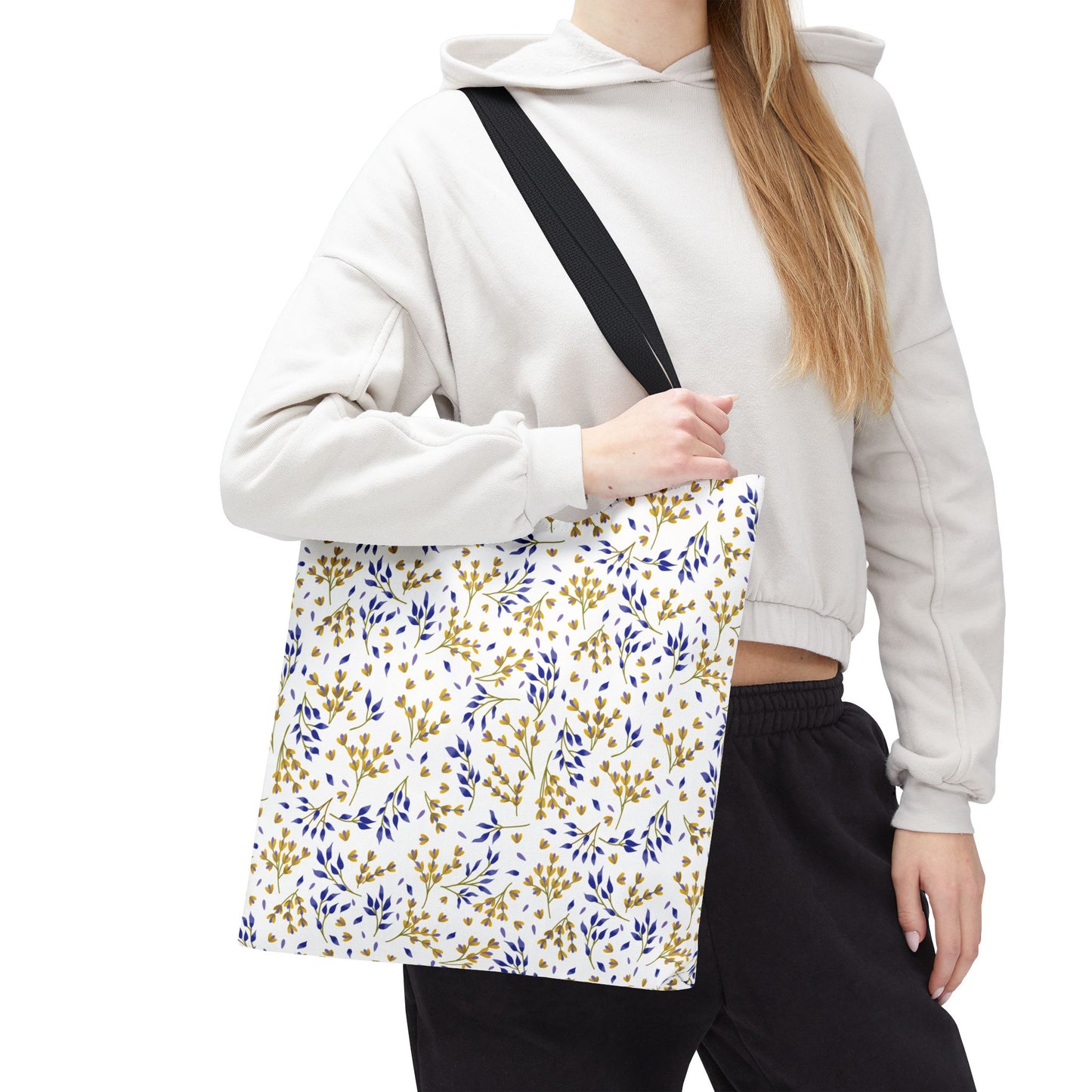 Person wearing a light-colored hoodie and dark pants carries the Wild Herb Garden Tote Bag, featuring gold-yellow branches and blue leaves, perfect for summer outings. The tote has a durable, white polyester body.