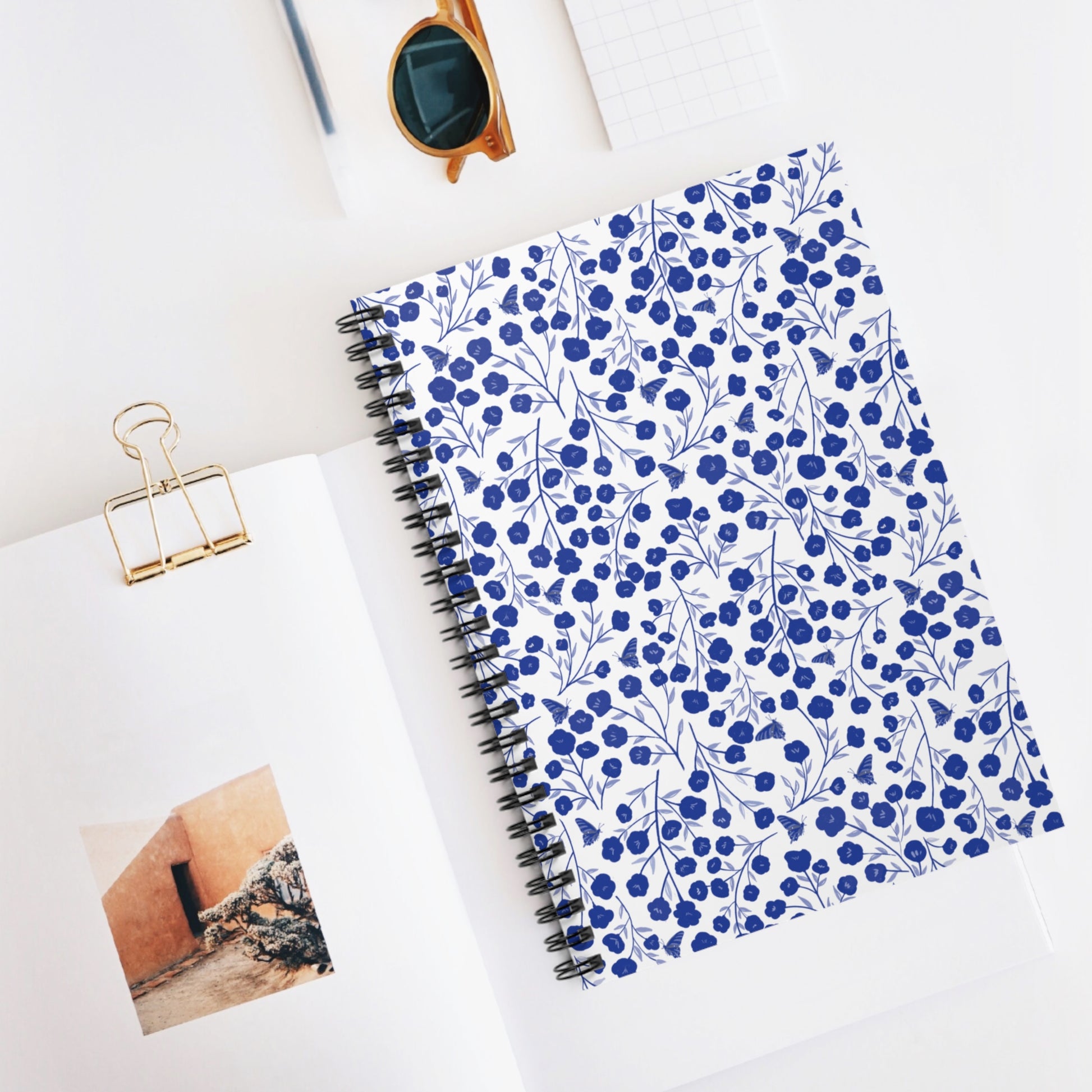 Floral Spiral Notebook with blue flowers and leafy patterns, neatly styled with an open book and sunglasses, epitomizes FireBrook Designs’ blend of style and functionality for creative expression and organization.