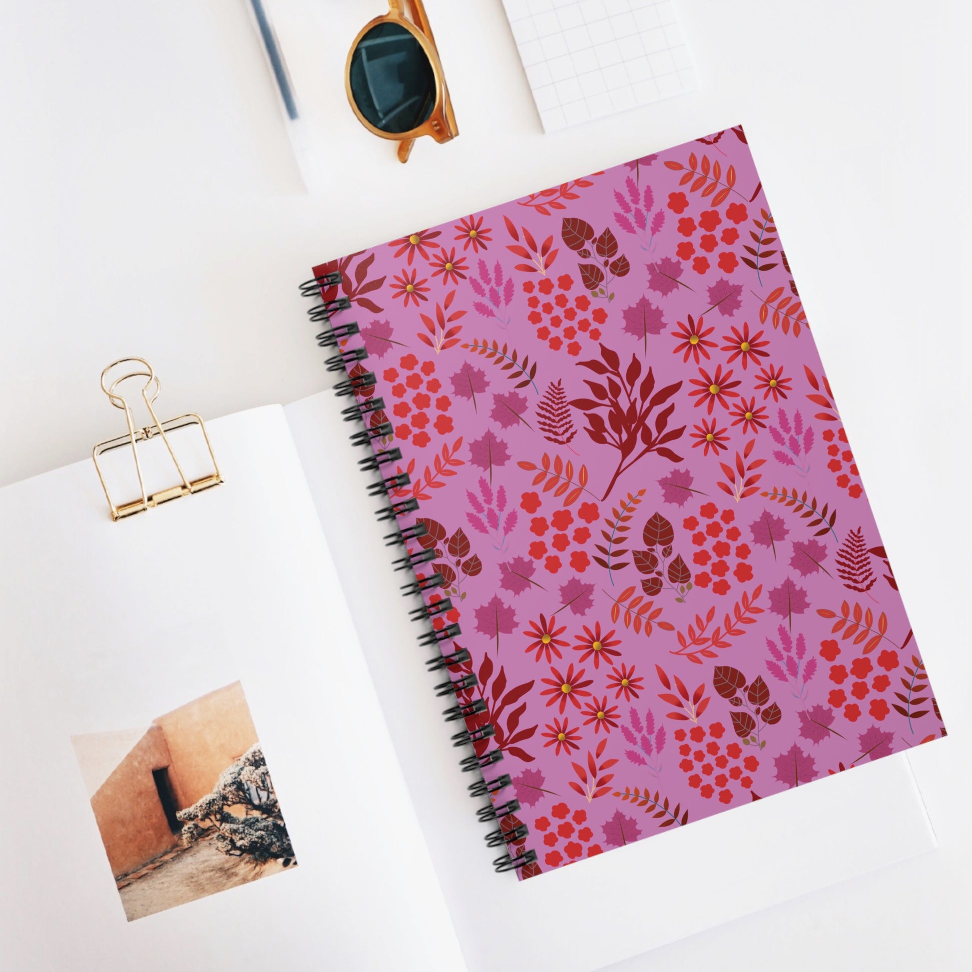 Floral Spiral Notebook with vibrant floral cover, placed diagonally on a white surface, exemplifies FireBrook Designs' uplifting style. A gold binder-clip holds an open book beneath, alongside tortoiseshell sunglasses.