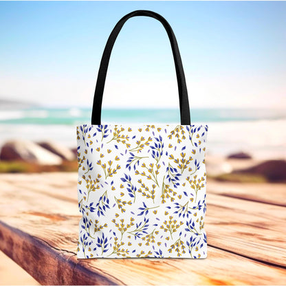 Floral Tote Bag with black handles, featuring blue and mustard-yellow sprigs on white, sits on a wooden surface near a beach, embodying FireBrook Designs' joyful and stylish essence, perfect for summer outings.