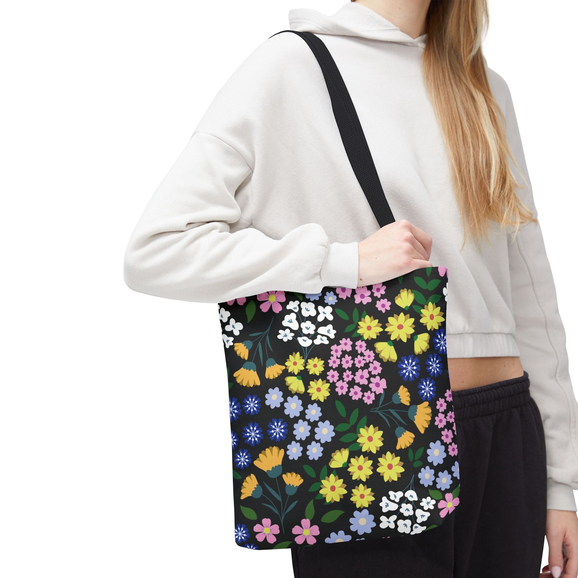 Person wearing light hoodie and dark pants carries the Floral Tote Bag from FireBrook Designs, featuring vibrant blossoms and green leaves on black, highlighting durability and style for joyful outings.