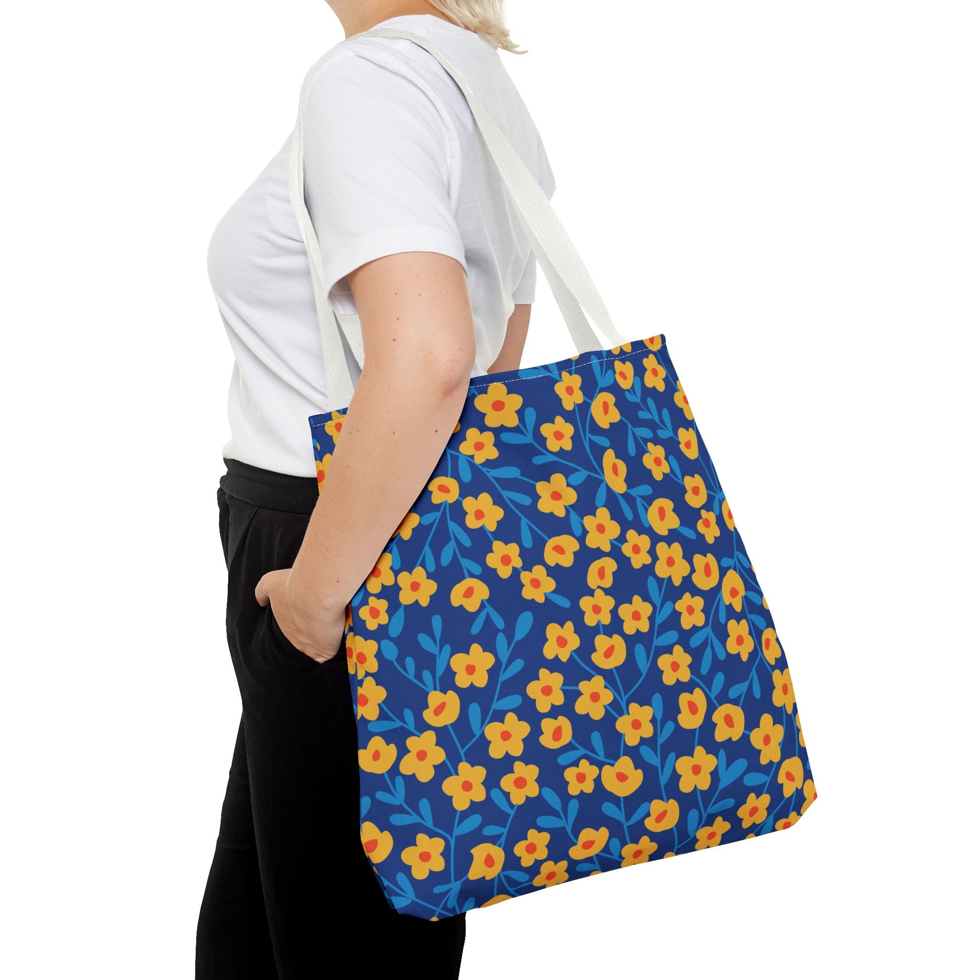 Person in white T-shirt carries the Cobalt Poppy Field Tote Bag, featuring yellow flowers with red centers and blue leaves on dark blue, showcasing FireBrook Designs' cheerful and durable style.