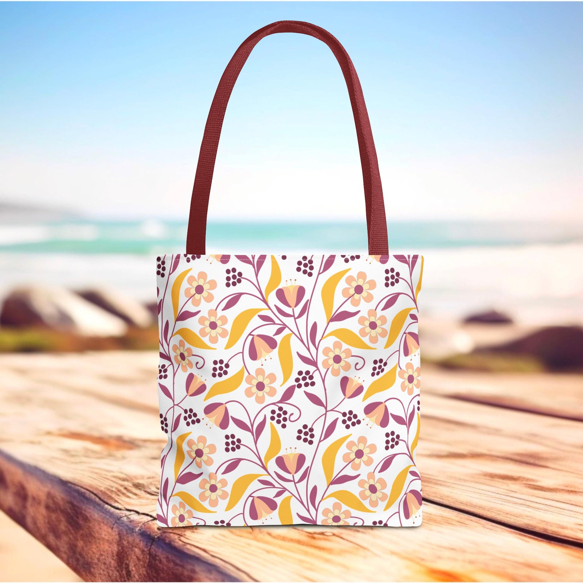 Floral Tote Bag on wooden deck with beach view; features a vibrant pink, mauve, and golden yellow floral pattern, ideal for summer outings. Durable polyester with reinforced handles, designed for style and positivity.