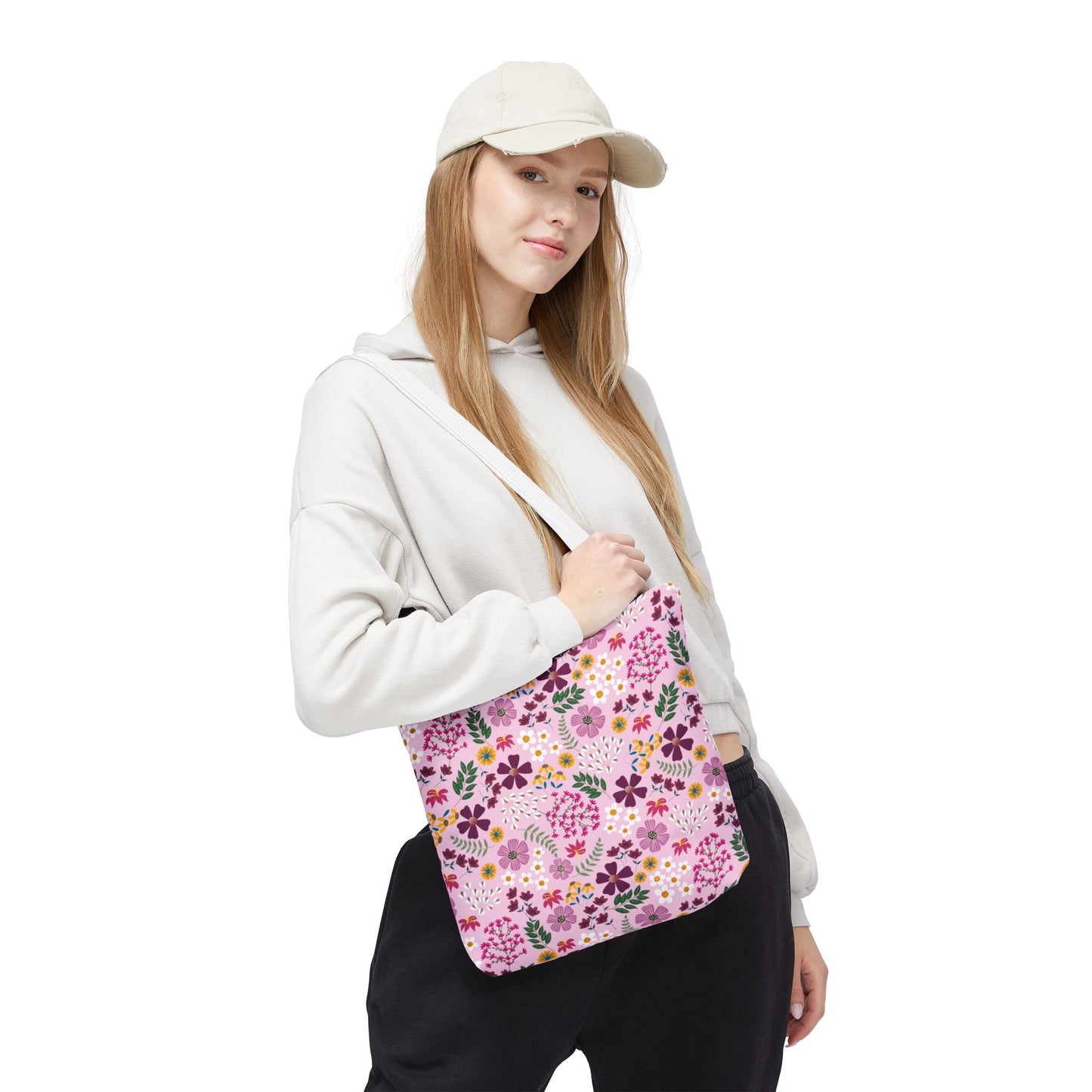 Young woman wearing a hoodie and cap holds the Rose Garden Blush Tote Bag, showcasing its floral pattern and casual style. Ideal for daily use, this bag complements FireBrook Designs' uplifting and practical ethos.