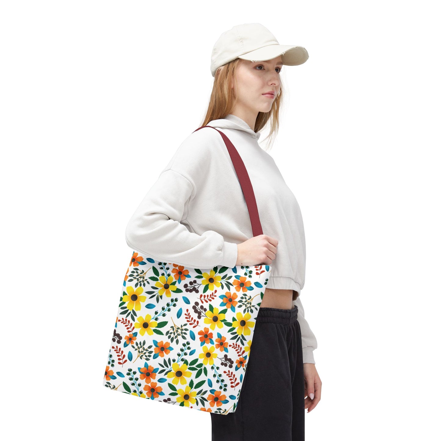 A young woman showcases the Sunny Meadow Mix Tote Bag, featuring a vibrant floral pattern with a burgundy strap, ideal for summer outings. She's styled in casual streetwear with a hoodie and joggers.