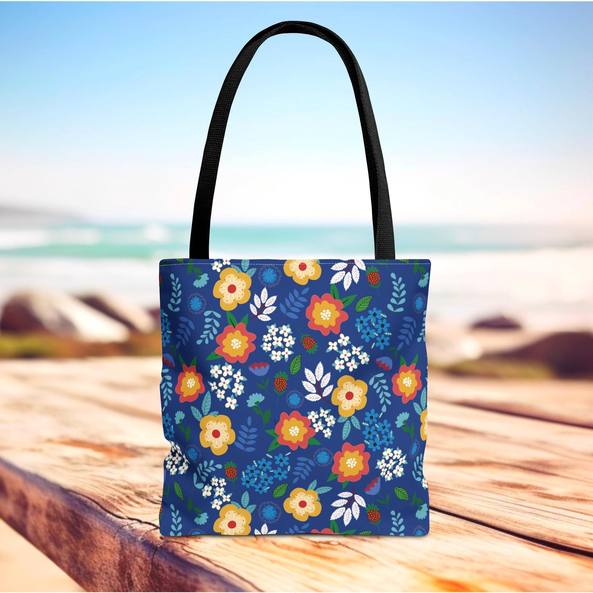 Cornflower Patch Tote Bag with black handles and a vibrant floral pattern on a deep-blue body, showcased on a wooden surface with a beach backdrop. Ideal for joyful summer outings.