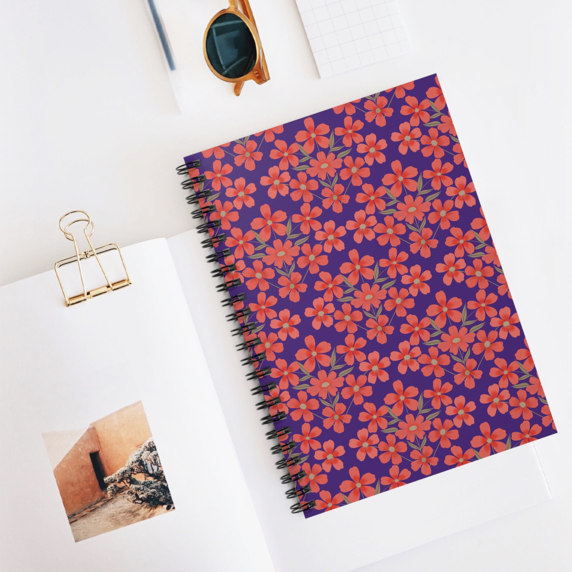 Floral Spiral Notebook: Overhead view showcasing a vibrant purple notebook with red and coral floral patterns, ideal for creative note-taking, surrounded by sunglasses and graph paper on a white surface.