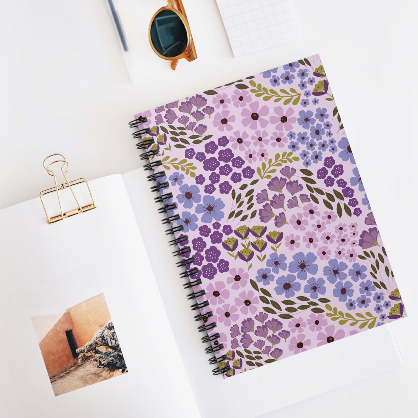 Floral Spiral Notebook with purple and lilac flower pattern, central on a white desk. Nearby, a gold binder clip and sunglasses add stylish accents, reflecting FireBrook Designs' uplifting, creative ethos.