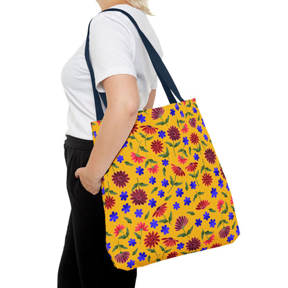 Person carrying the Golden Wildflower Tote Bag with navy straps, featuring a vibrant yellow body adorned with red and maroon daisies, small blue flowers, and green leaves against a floral pattern.