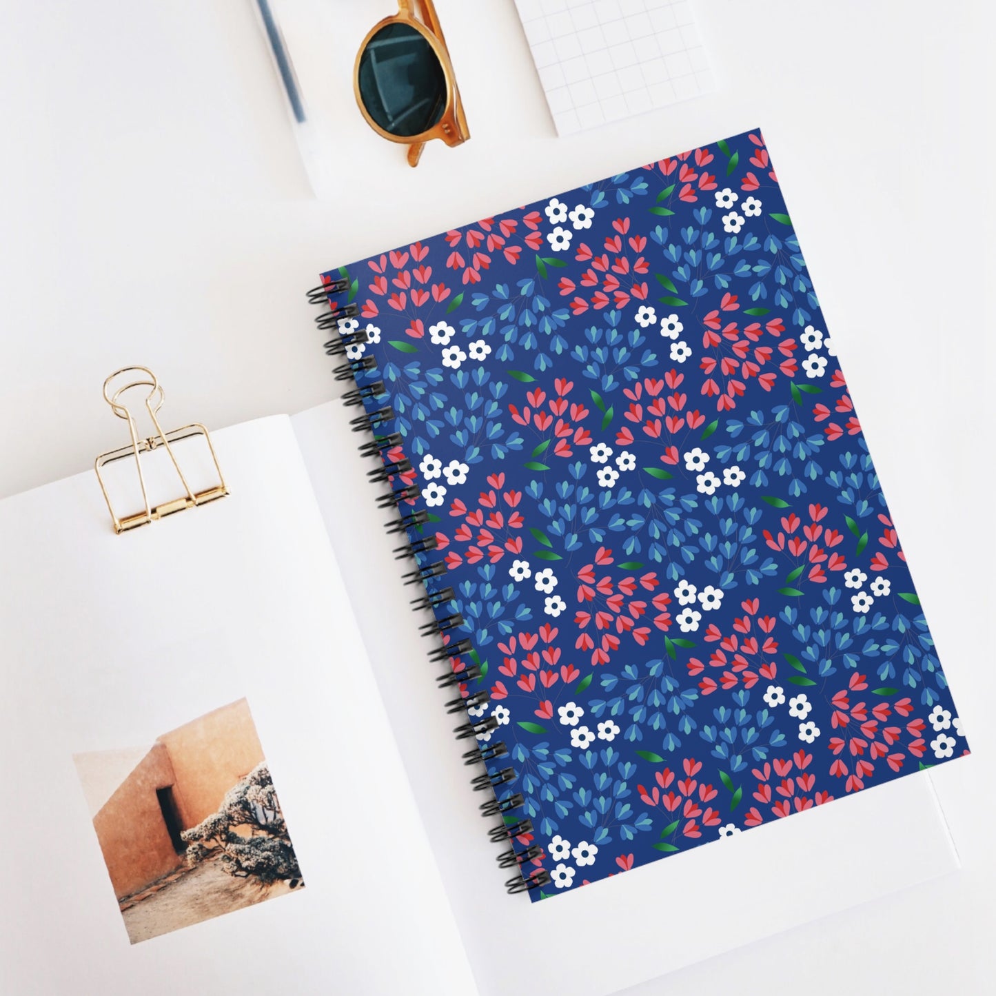 Floral Spiral Notebook with vibrant floral motifs, centered in a flat-lay scene with a gold binder clip, sunglasses, and graph paper, reflecting FireBrook Essentials' ethos of stylish, joyful functionality.