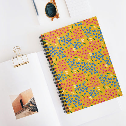Floral Spiral Notebook with a mustard-yellow cover, adorned with vibrant floral patterns, lies on an open magazine. Nearby, amber-framed sunglasses add a stylish touch. Perfect for creative note-taking.