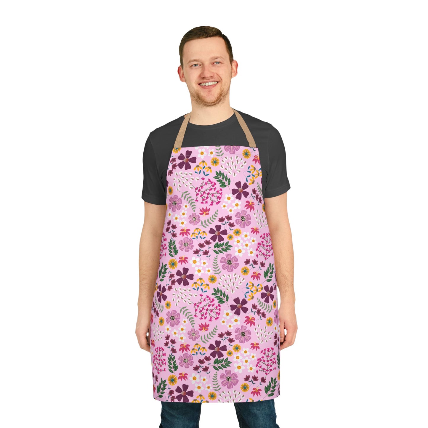 Young man in a dark-gray T-shirt and jeans wears a FireBrook Designs Floral Apron, featuring a pink base with vibrant floral patterns, showcasing style and functionality for cooking or crafting.