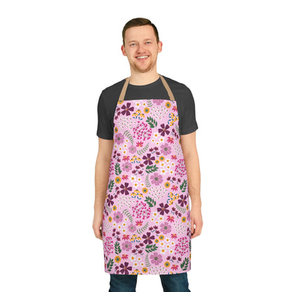 Young man in a dark-gray T-shirt and jeans wears a FireBrook Designs Floral Apron, featuring a pink base with vibrant floral patterns, showcasing style and functionality for cooking or crafting.