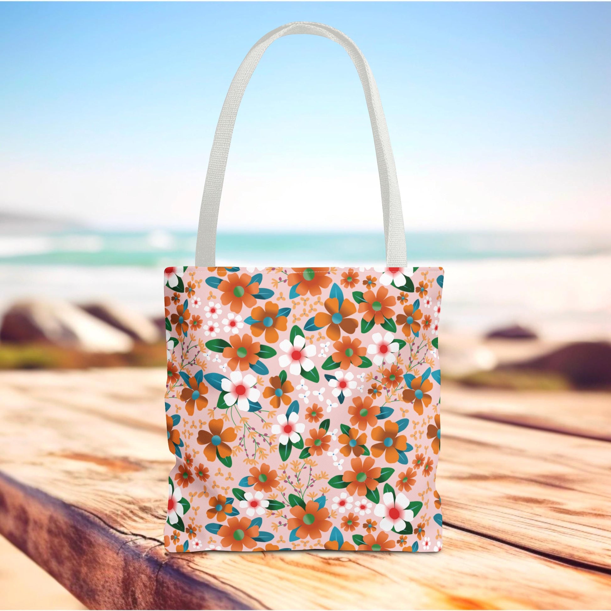 Peach Poppy Grove Tote Bag featuring vibrant orange and white floral patterns with green and teal leaves on a pale pink backdrop, perfect for summer outings. The bag sits on a wooden surface near the beach.