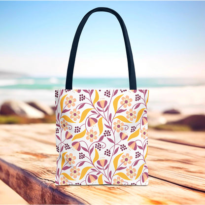 Floral Tote Bag with dark straps features a cheerful floral motif in pinks, mustard yellow, and burgundy, ideal for summer outings. Displayed on a wooden surface with ocean background.