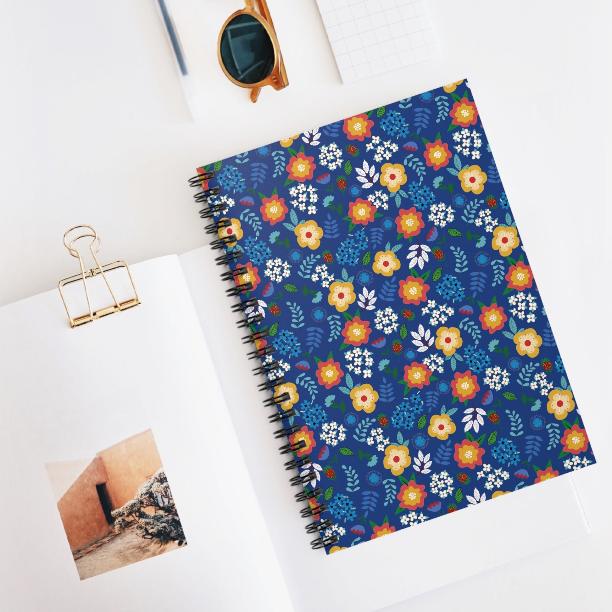 Floral Spiral Notebook with vibrant flower pattern on dark blue cover, central to a white desk scene, embodies FireBrook Designs' joyful, uplifting style, enhancing creativity for students and professionals alike.
