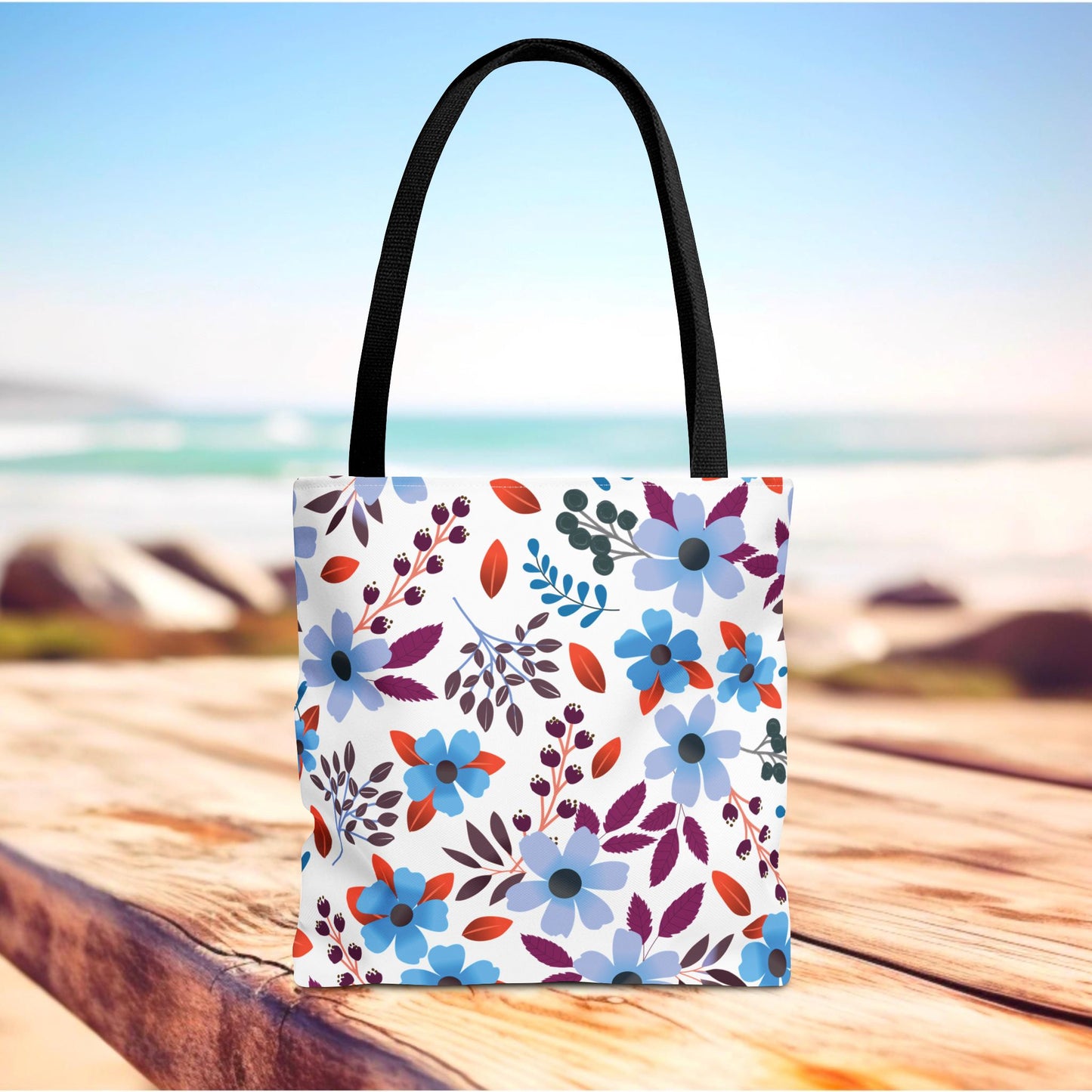 Floral Tote Bag with vibrant blues and purples, stands on a wooden plank near a beach. Designed for durability with polyester fabric and reinforced handles, perfect for joyful summer outings.