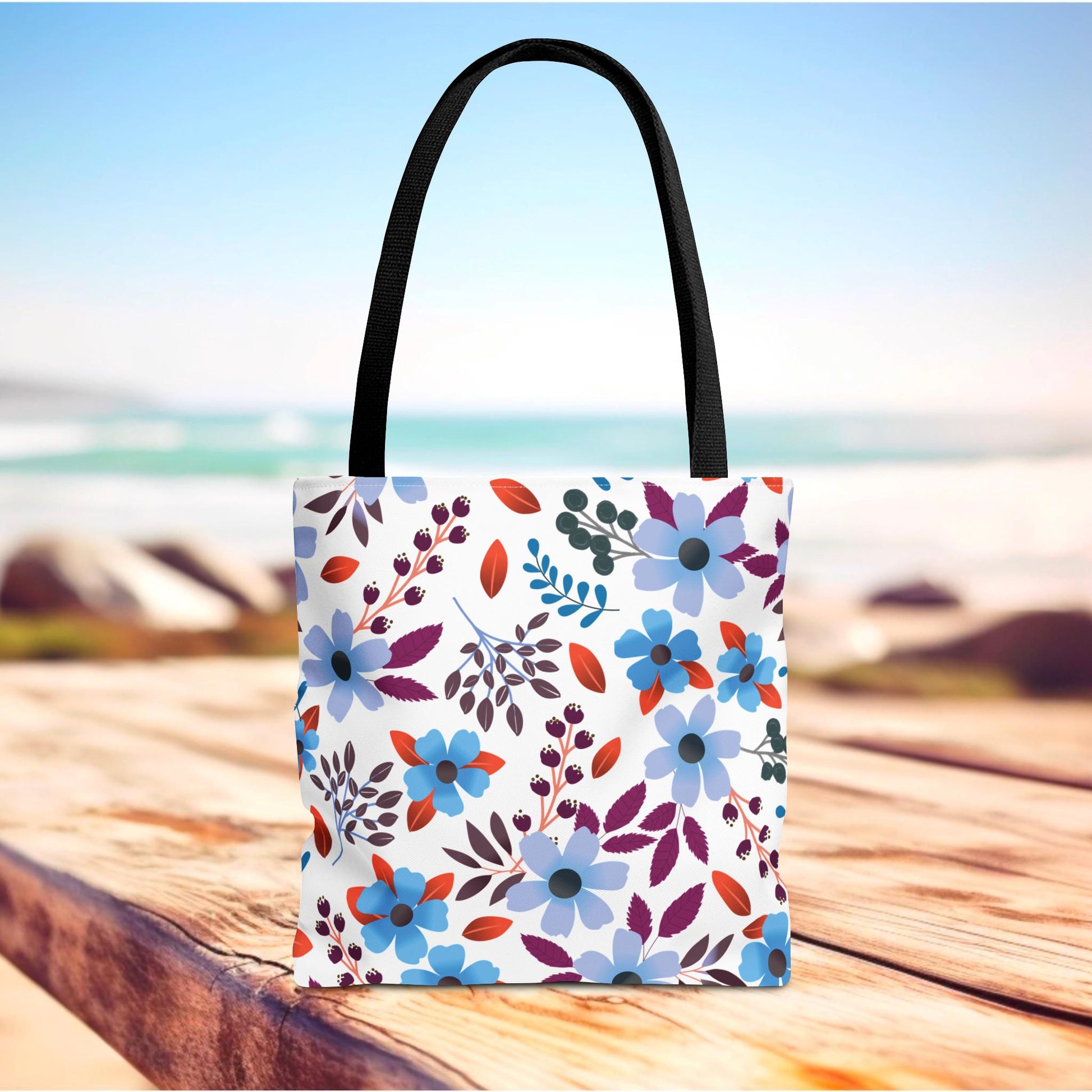 Floral Tote Bag with vibrant blues and purples, stands on a wooden plank near a beach. Designed for durability with polyester fabric and reinforced handles, perfect for joyful summer outings.