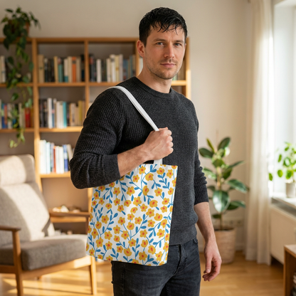 Man in a living room wears a dark sweater and jeans, showcasing the Buttercup Cream Tote Bag with a vibrant floral pattern, embodying FireBrook Essentials' joyful, uplifting design ethos.