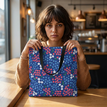 Young woman at café table holds the Berry Blue Festival Tote Bag, featuring a colorful floral print with small blossoms and leaves, embodying FireBrook Designs' cheerful, stylish essence.