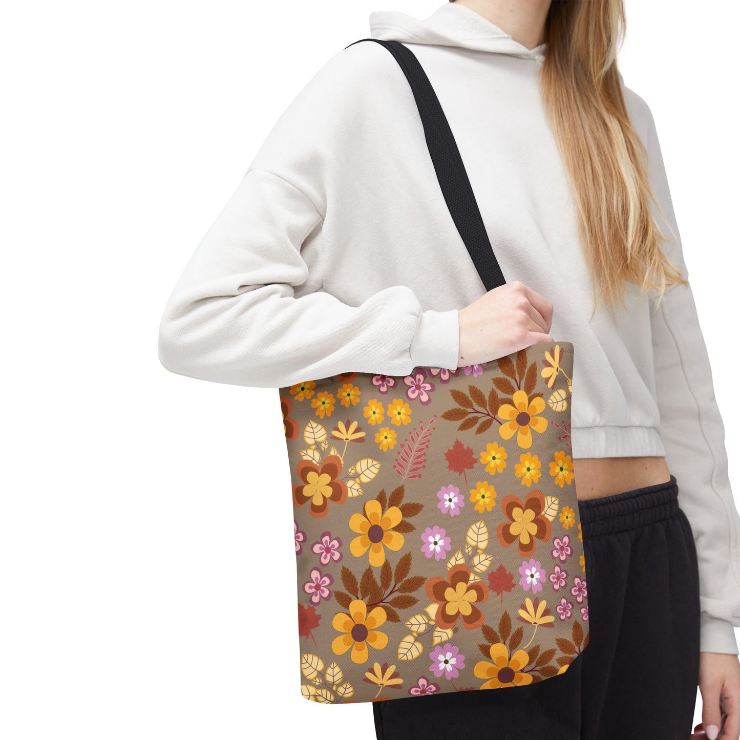 The Harvest Brown Bloom Tote Bag features a fall-inspired floral pattern with orange, yellow, and reddish-brown flowers on a brown background. It's being held by someone wearing a light-colored cropped hoodie.