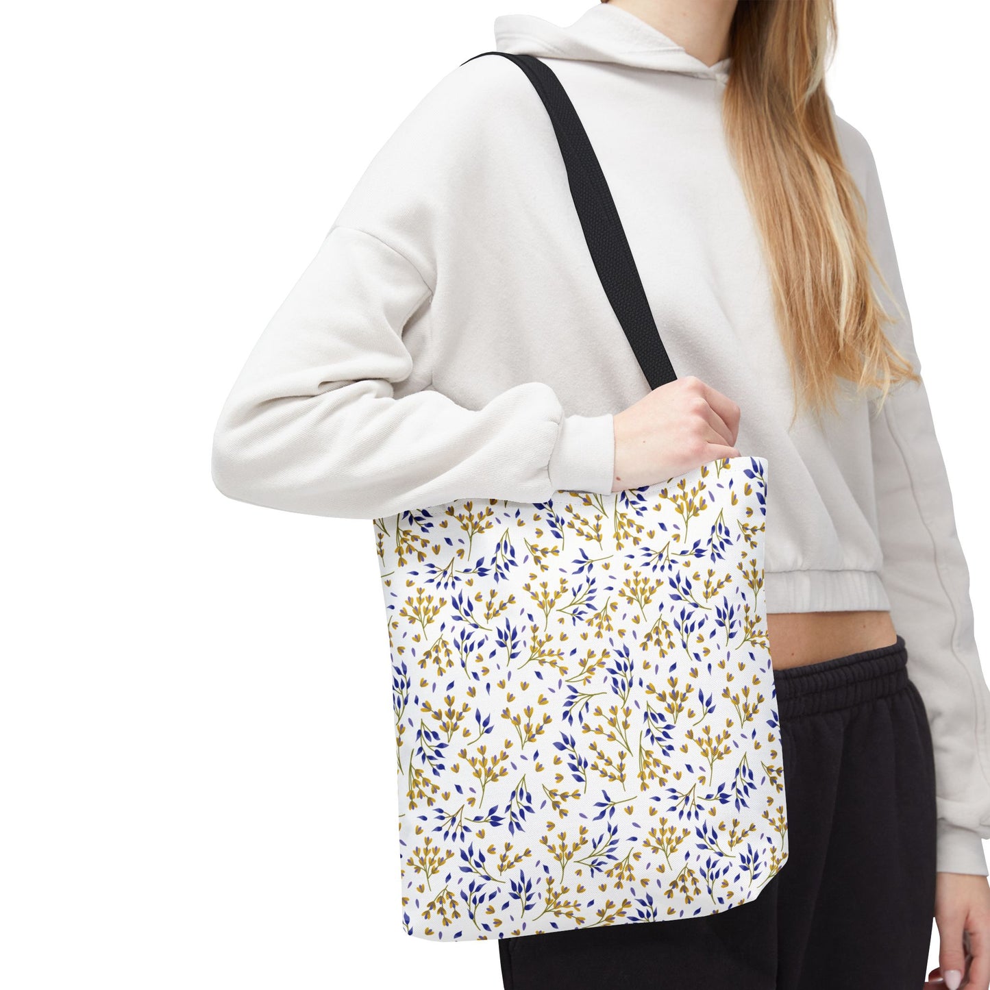 Person wearing cropped hoodie holds the Wild Herb Garden Tote Bag with a botanical print in mustard yellow and deep blue, featuring black handles, perfect for cheerful, stylish outings.