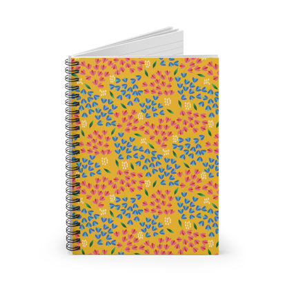 Floral Spiral Notebook with a vibrant mustard-yellow cover featuring a pink, blue, and green floral motif, standing upright. Its open pages reveal lined paper, perfect for creative note-taking or journaling.