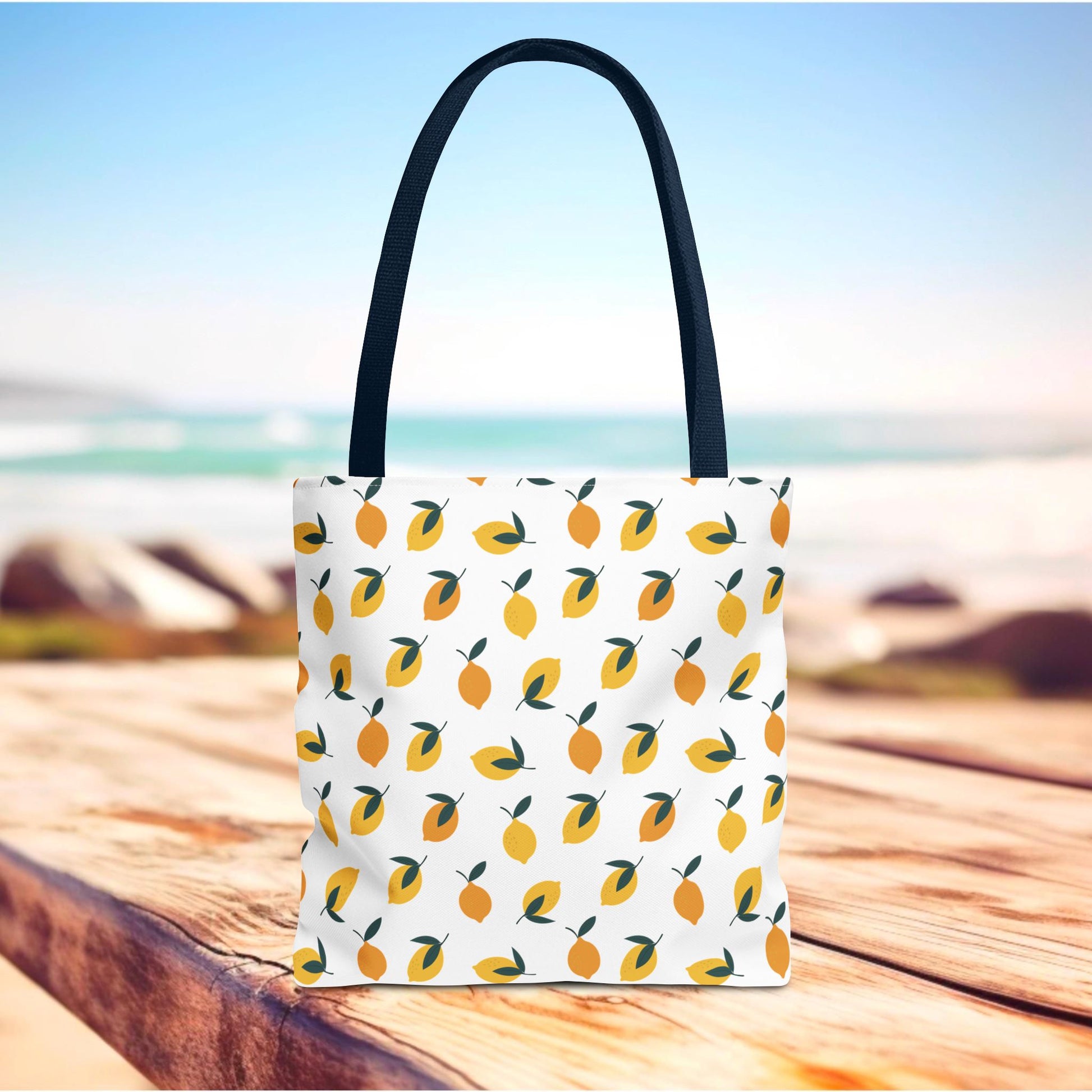 The Lemon Drop Toss Tote Bag, adorned with a lively lemon pattern, sits on a wooden surface against a beach backdrop, showcasing its vibrant, cheerful design ideal for summer outings.