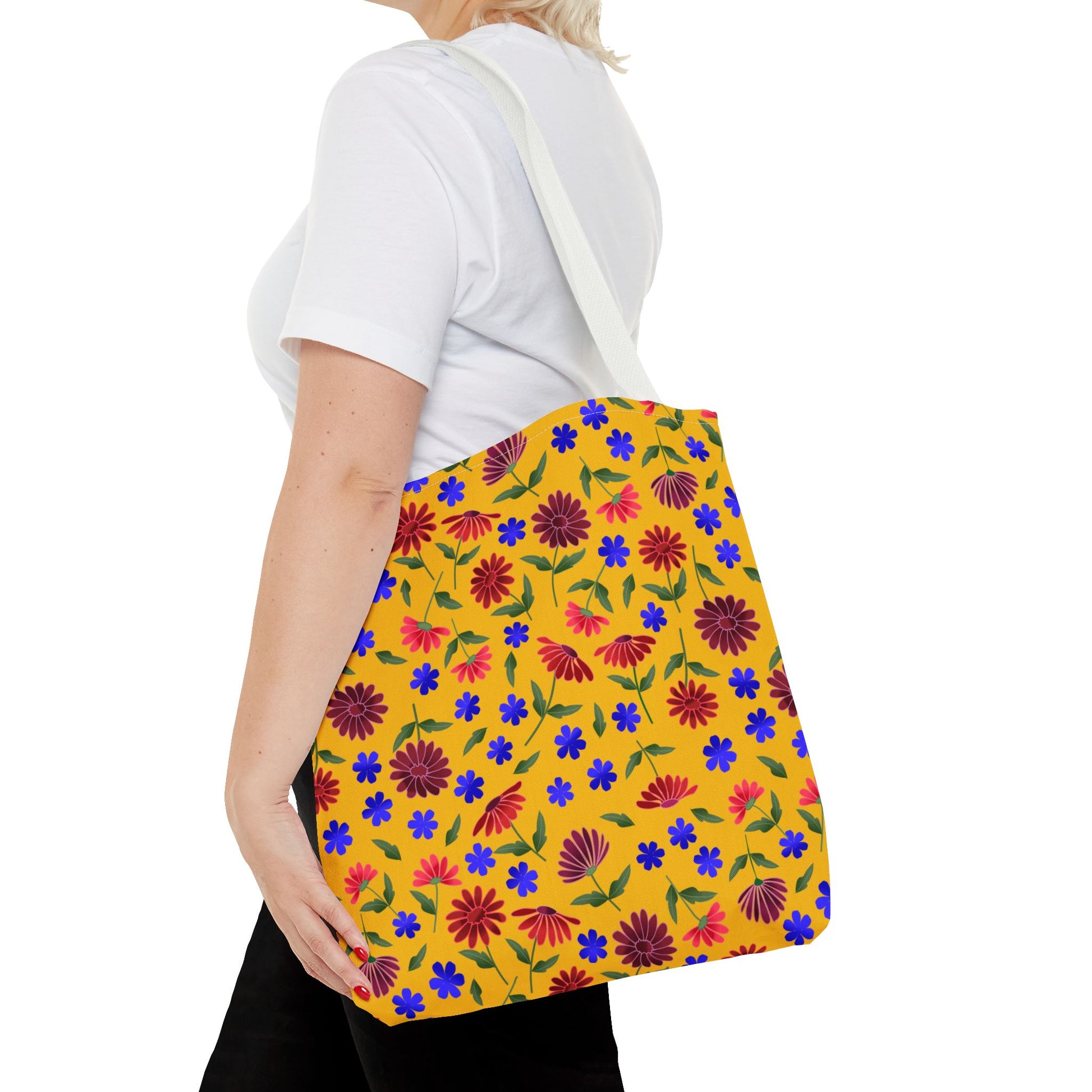 Golden Wildflower Tote Bag features a mustard-yellow backdrop with red daisies, green leaves, and blue flowers. Black straps enhance its durability and style, perfect for everyday use.