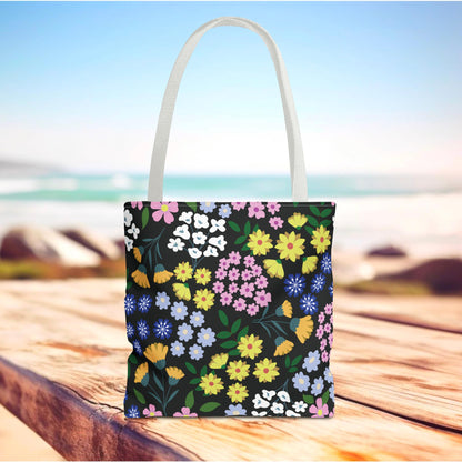 Floral Tote Bag with vibrant blossoms and green leaves on a black body, showcased on a sunlit wooden surface. Perfect for summer outings, reflecting FireBrook Designs' cheerful, uplifting aesthetic.