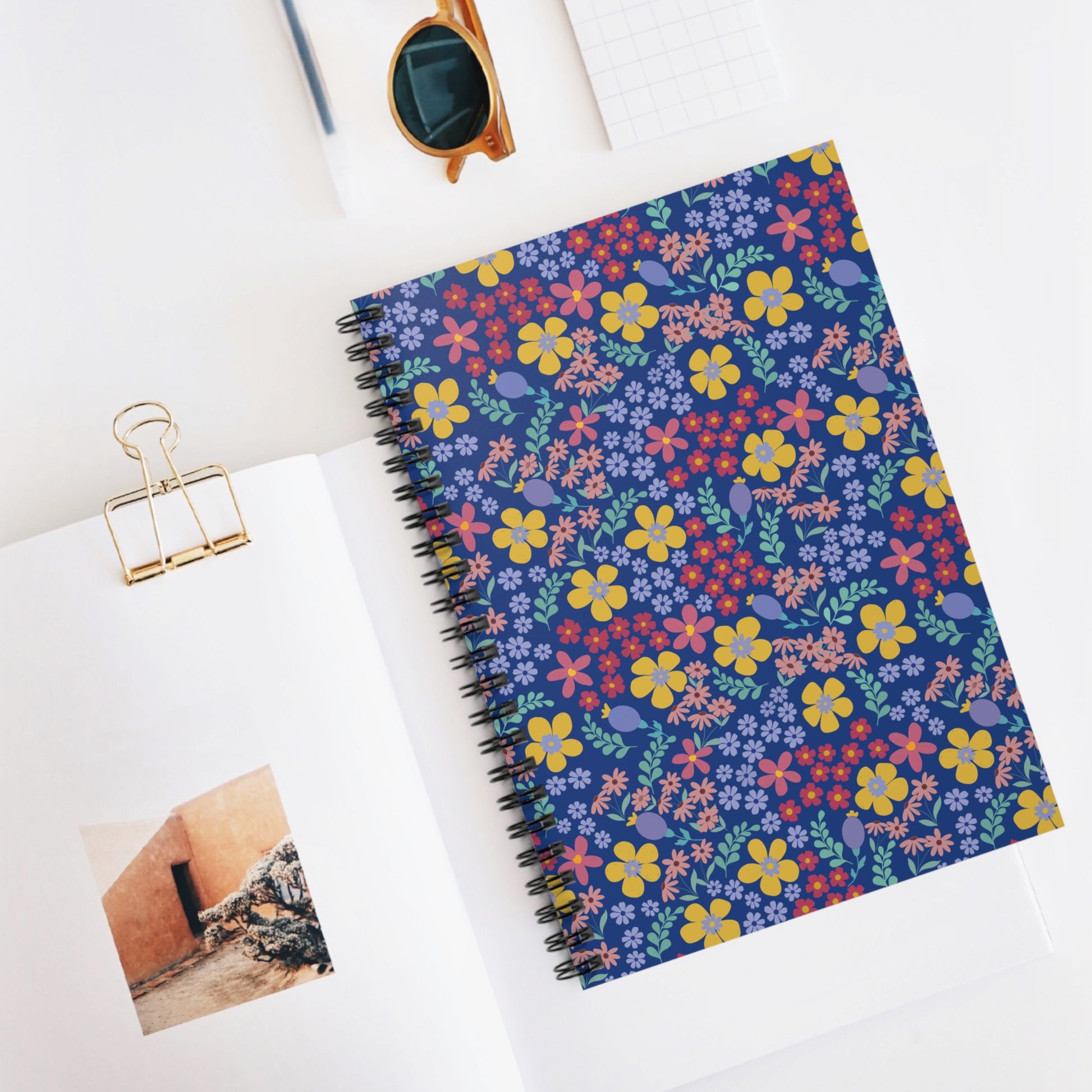 Floral Spiral Notebook with colorful flowers on a dark blue cover, paired with round sunglasses and a small gold binder clip, embodies FireBrook Essentials' uplifting, creative spirit for joyful note-taking.
