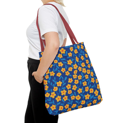 A person carries the Cobalt Poppy Field Tote Bag over their shoulder, showcasing its vibrant yellow-orange floral pattern on a deep blue background, with dark red straps, ideal for summer adventures.