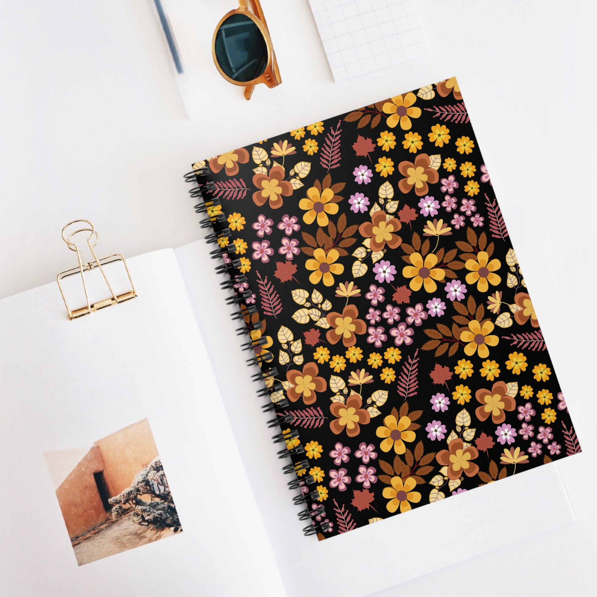 Floral Spiral Notebook with warm-toned floral pattern against black, adorned with sleek metal spiral binding, rests on a white desktop alongside a magazine, inviting creativity and organization.