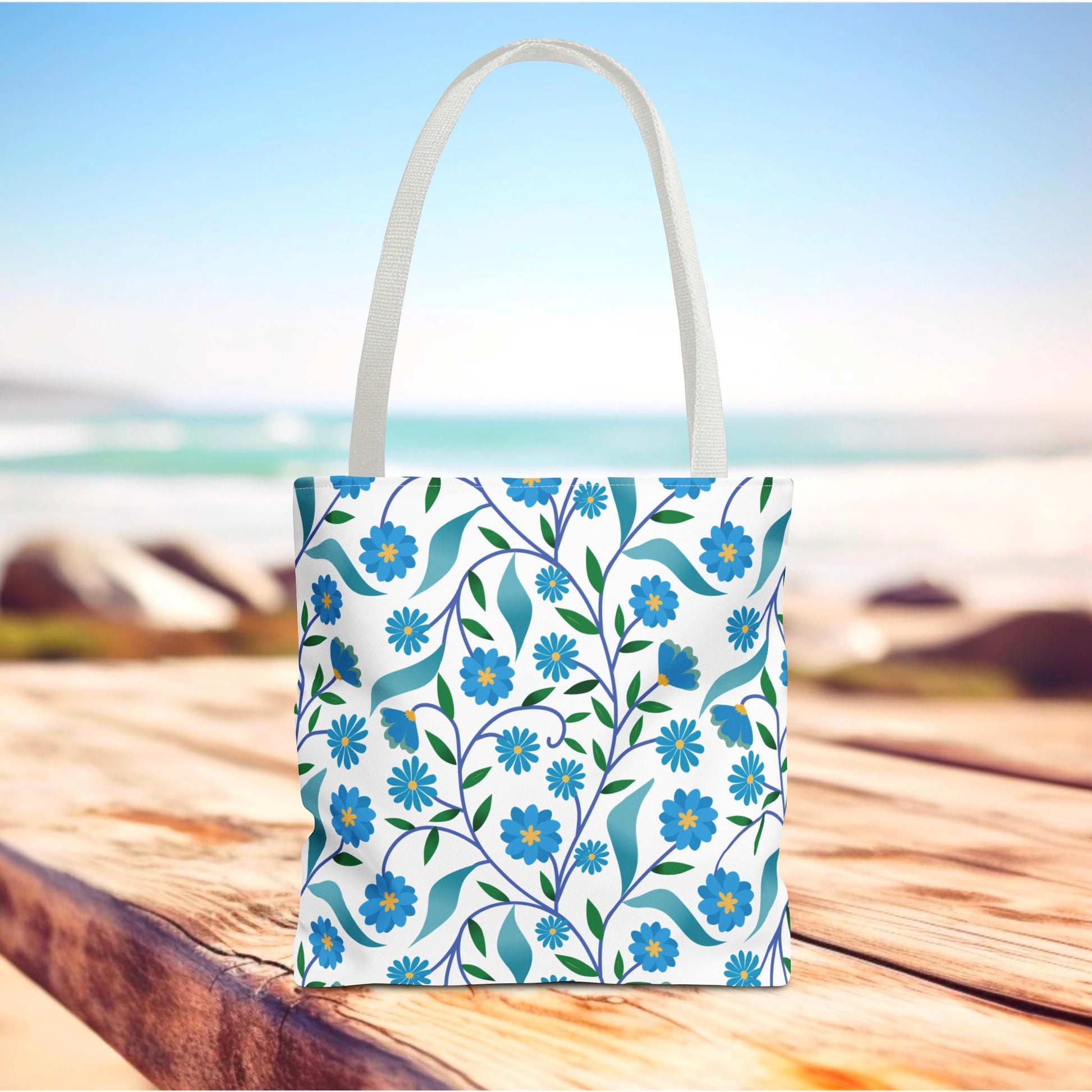 Blue Floral Tote Bag with white straps, featuring a vibrant pattern of blue flowers, green leaves, and curling stems. Styled outdoors on wood, reflecting FireBrook Essentials' joyful and uplifting design ethos.