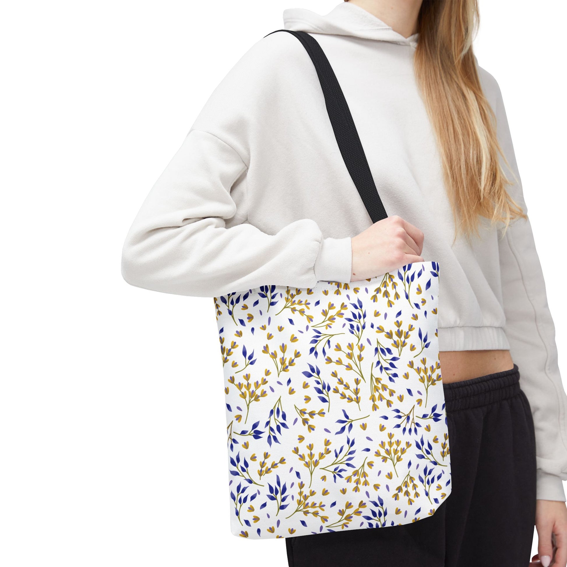 Floral Tote Bag featuring a small, repeating botanical pattern in yellow-gold and deep blue, held by a person wearing a light hoodie. Ideal for summer outings with durable polyester and reinforced black handles.